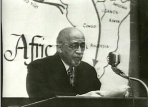 W.E.B. DuBois sits in front of a microphone with a map of Africa behind him