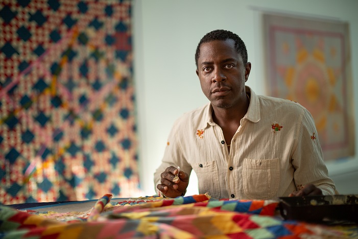 Sanford Biggers sits with a quilt on the table in front of him and additional quilts hung on the wall.